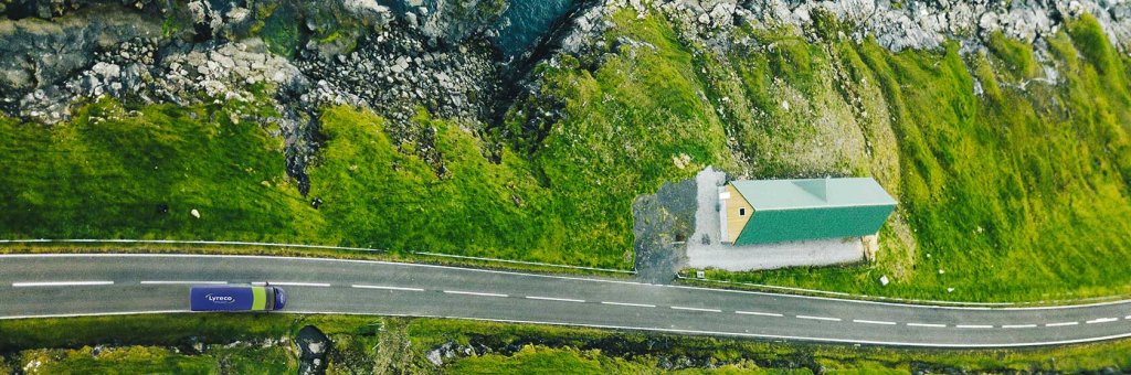 Luftaufnahme einer grünen Landschaft und einer rauen Küstenstraße, auf der ein Lyreco-Auslieferfahrzeug fährt und an der ein Haus steht.