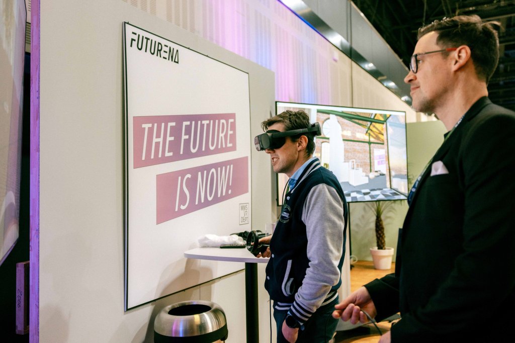 Robin, Interior Design Expert von MYWORKSPACE by Lyreco, rechts im Bild im schwarzen Anzug mit Brille zeigt Xavier Etienne, Managing Director Europe von Lyreco, der mit VR-Brille auf dem Kopf und Fernbedienung in der Hand auf einen Bildschirm schaut, eine virtuelle Planung. (Bildquelle: Spieker Fotografie).