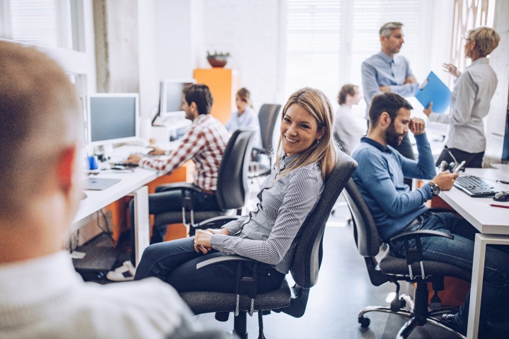 Eine Frau sitzt in einem Büro auf einem Bürostuhl und schaut lächelnd zu einem Kollegen. Das Büro ist mit Mitarbeitern gefüllt, die arbeiten.