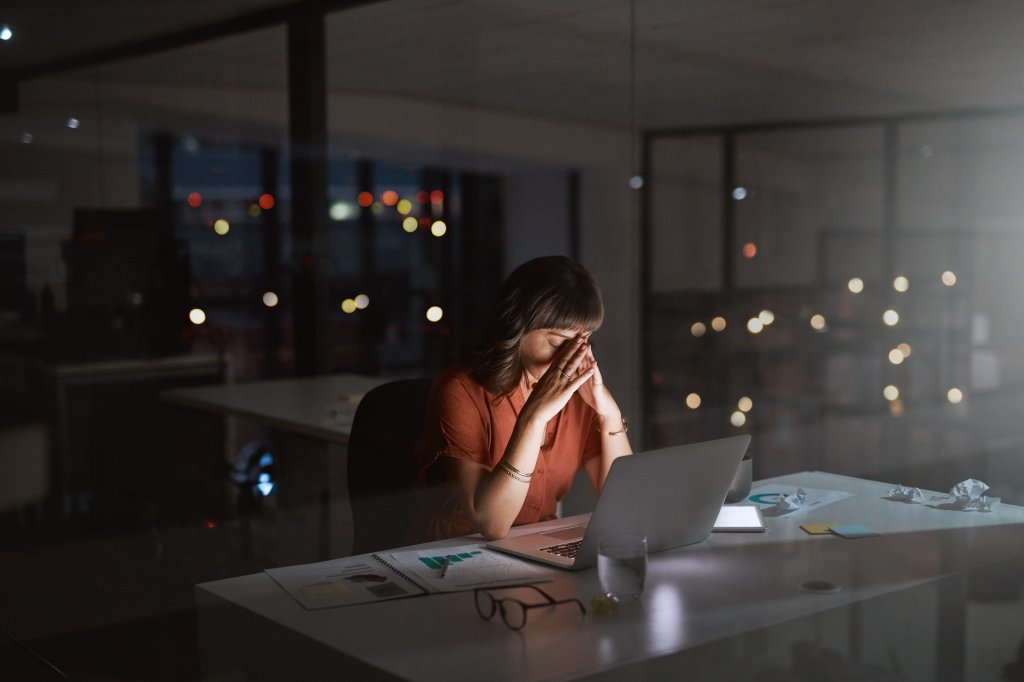 Aufnahme einer jungen Geschäftsfrau, die gestresst aussieht, während sie im Dunkeln in einem Büro an einem Laptop arbeitet. Mit den richtigen Tipps von MYWORKSPACE by Lyreco können Sie diverse Produktivitätskiller im Büro vermeiden.
