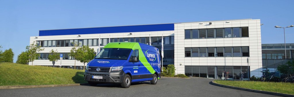 Im Vordergrund ein blaues Auslieferfahrzeug von Lyreco. Im Hintergrund ein weißes Bürogebäude, welches das Headquarter der Lyreco Deutschland GmbH in Barsinghausen ist.