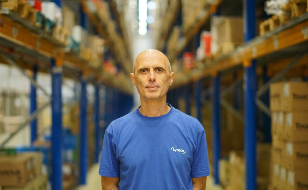 Logistik-Mitarbeiter von Lyreco im blauen T-Shirt mit Lyreco-Logo. Er steht zwischen den Hochregallagerplätzen.
